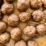 Baked meatballs resting on a parchment-lined sheet pan with browned tops and a metal spatula taking a few from the pan.