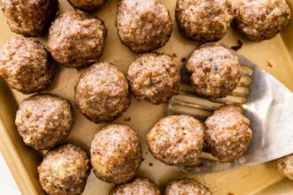 Baked meatballs resting on a parchment-lined sheet pan with browned tops and a metal spatula taking a few from the pan.