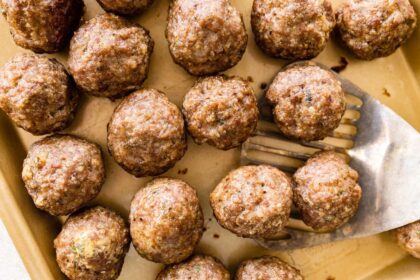 Baked meatballs resting on a parchment-lined sheet pan with browned tops and a metal spatula taking a few from the pan.