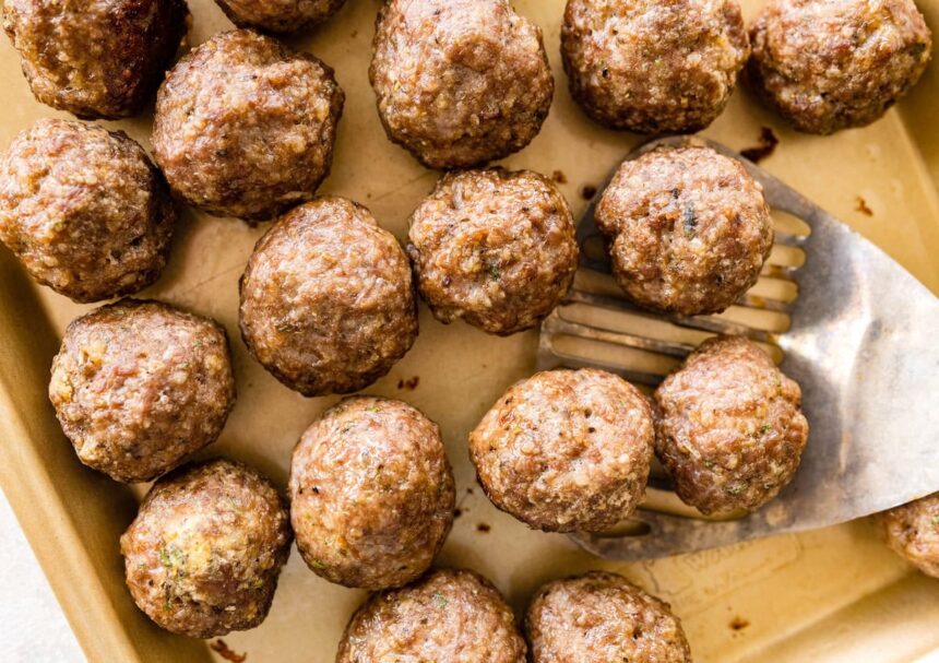 Baked meatballs resting on a parchment-lined sheet pan with browned tops and a metal spatula taking a few from the pan.