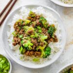 Ground beef and broccoli served on a bed of white rice, topped with sesame seeds and green onions. Wooden chopsticks are next to the bowl.