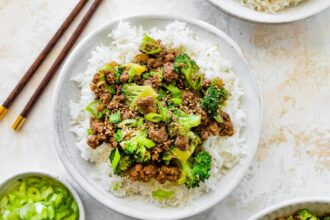 Ground beef and broccoli served on a bed of white rice, topped with sesame seeds and green onions. Wooden chopsticks are next to the bowl.