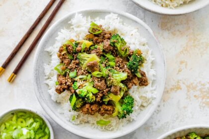Ground beef and broccoli served on a bed of white rice, topped with sesame seeds and green onions. Wooden chopsticks are next to the bowl.