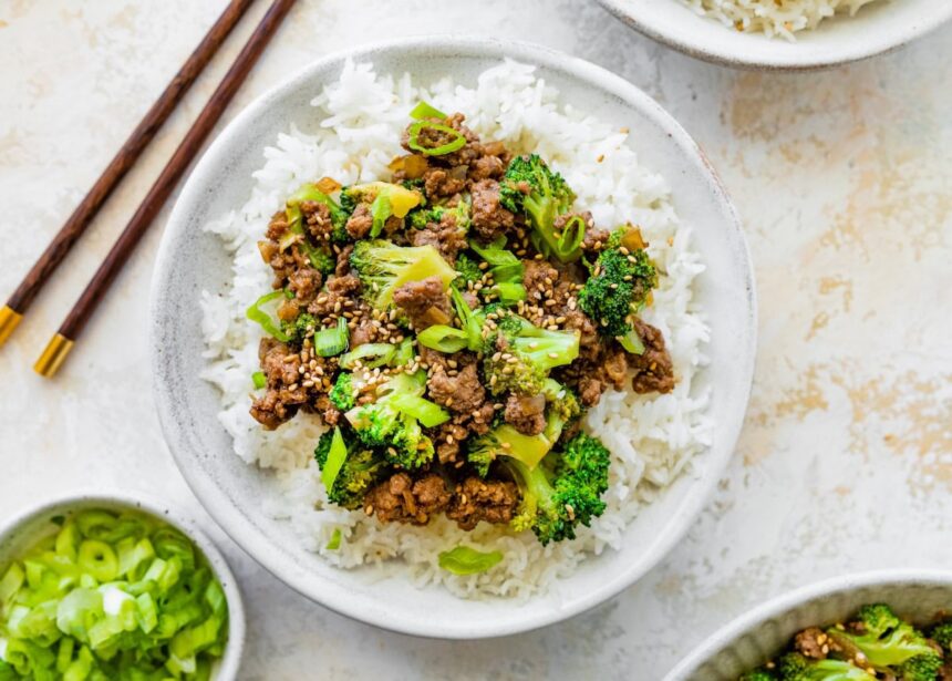 Ground beef and broccoli served on a bed of white rice, topped with sesame seeds and green onions. Wooden chopsticks are next to the bowl.