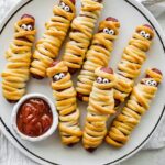 Plate of baked mummy hot dogs wrapped in crescent roll dough with candy eyes, served with ketchup for dipping.