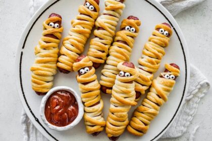 Plate of baked mummy hot dogs wrapped in crescent roll dough with candy eyes, served with ketchup for dipping.