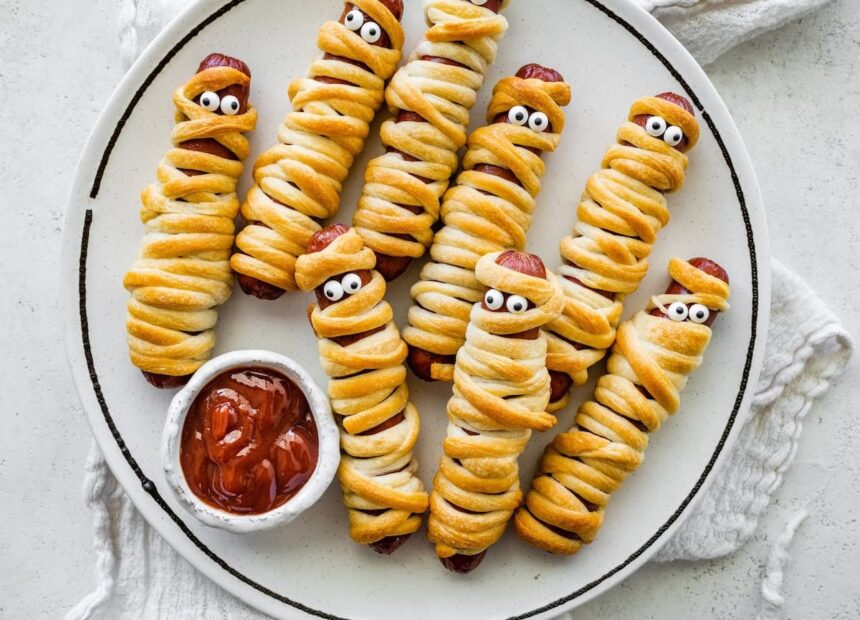 Plate of baked mummy hot dogs wrapped in crescent roll dough with candy eyes, served with ketchup for dipping.