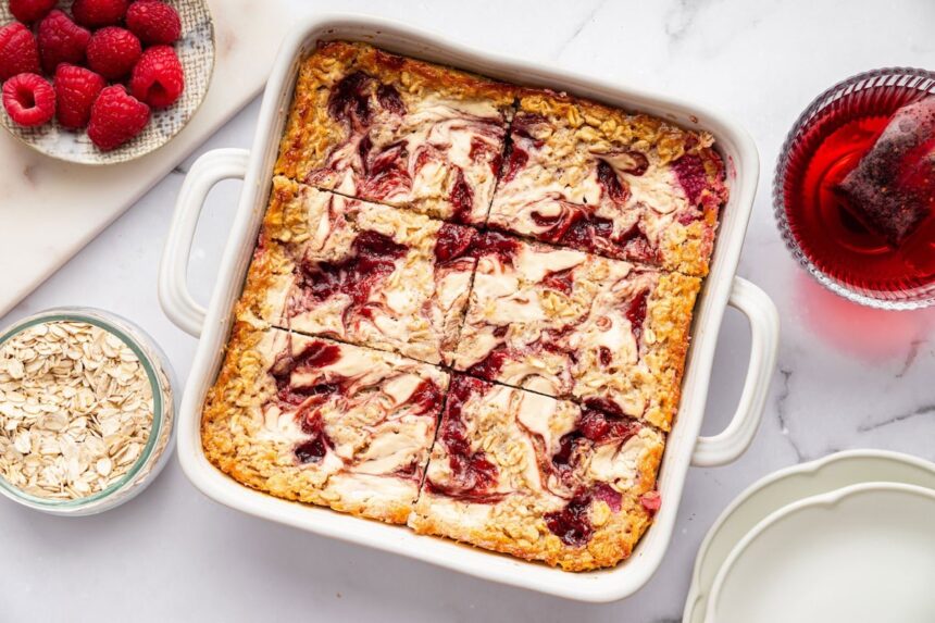 Baked oatmeal in a white baking dish showing creamy cottage cheese and raspberry jam swirls on top. Fresh raspberries and rolled oats are next to the dish.