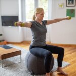 woman sitting down on a gray stability ball doing a dumbbell workout in living room