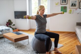 woman sitting down on a gray stability ball doing a dumbbell workout in living room