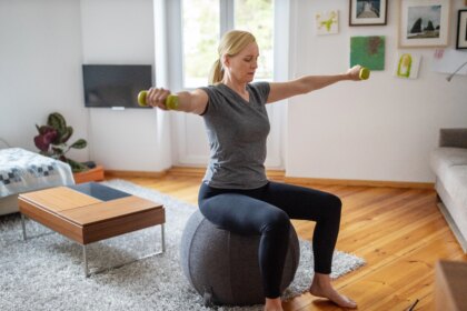 woman sitting down on a gray stability ball doing a dumbbell workout in living room