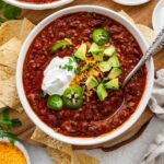 A bowl of slow cooked bean and beef chili topped with avocado, cheese, sour cream, and jalapeno slices.