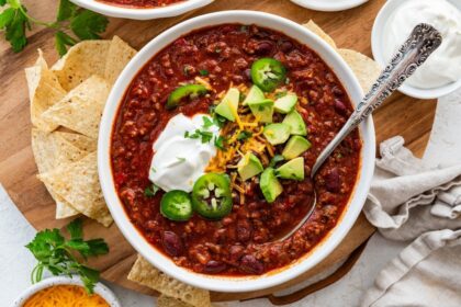 A bowl of slow cooked bean and beef chili topped with avocado, cheese, sour cream, and jalapeno slices.
