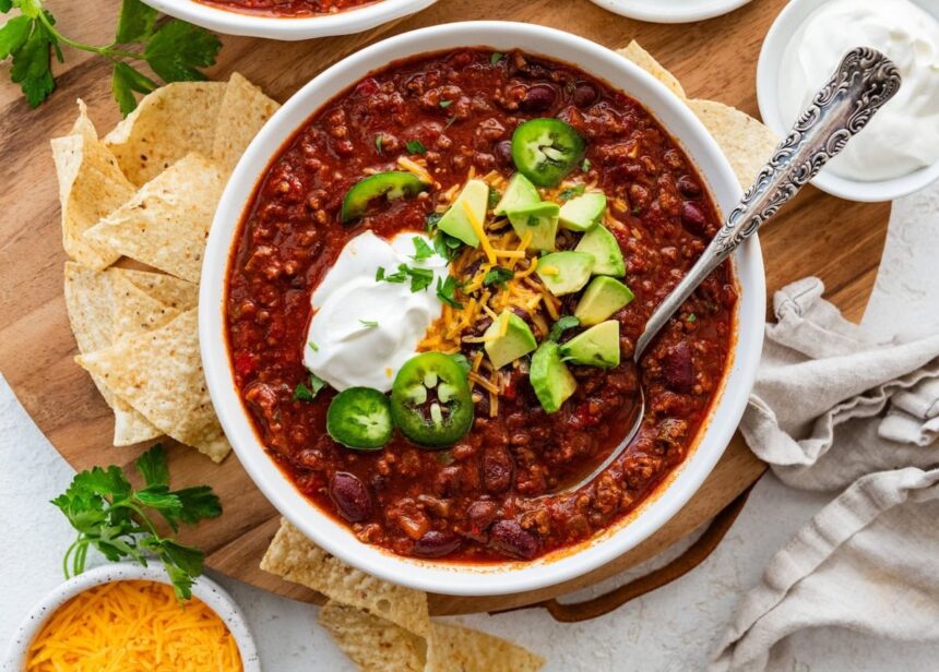 A bowl of slow cooked bean and beef chili topped with avocado, cheese, sour cream, and jalapeno slices.