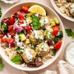 A serving of Greek turkey meatballs with brown rice, cucumber tomato salad, tzatziki, olives, and fresh herbs all in a bowl.