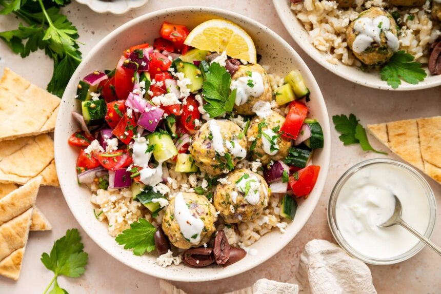 A serving of Greek turkey meatballs with brown rice, cucumber tomato salad, tzatziki, olives, and fresh herbs all in a bowl.