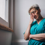 A healthcare worker in teal scrubs leans against a wall with their arms crossed and head bowed, lost in thought. The image captures a moment of contemplation and emotional fatigue.