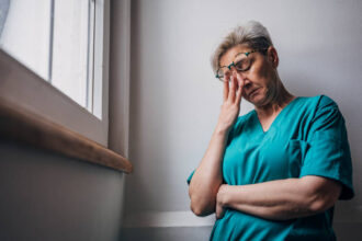 A healthcare worker in teal scrubs leans against a wall with their arms crossed and head bowed, lost in thought. The image captures a moment of contemplation and emotional fatigue.